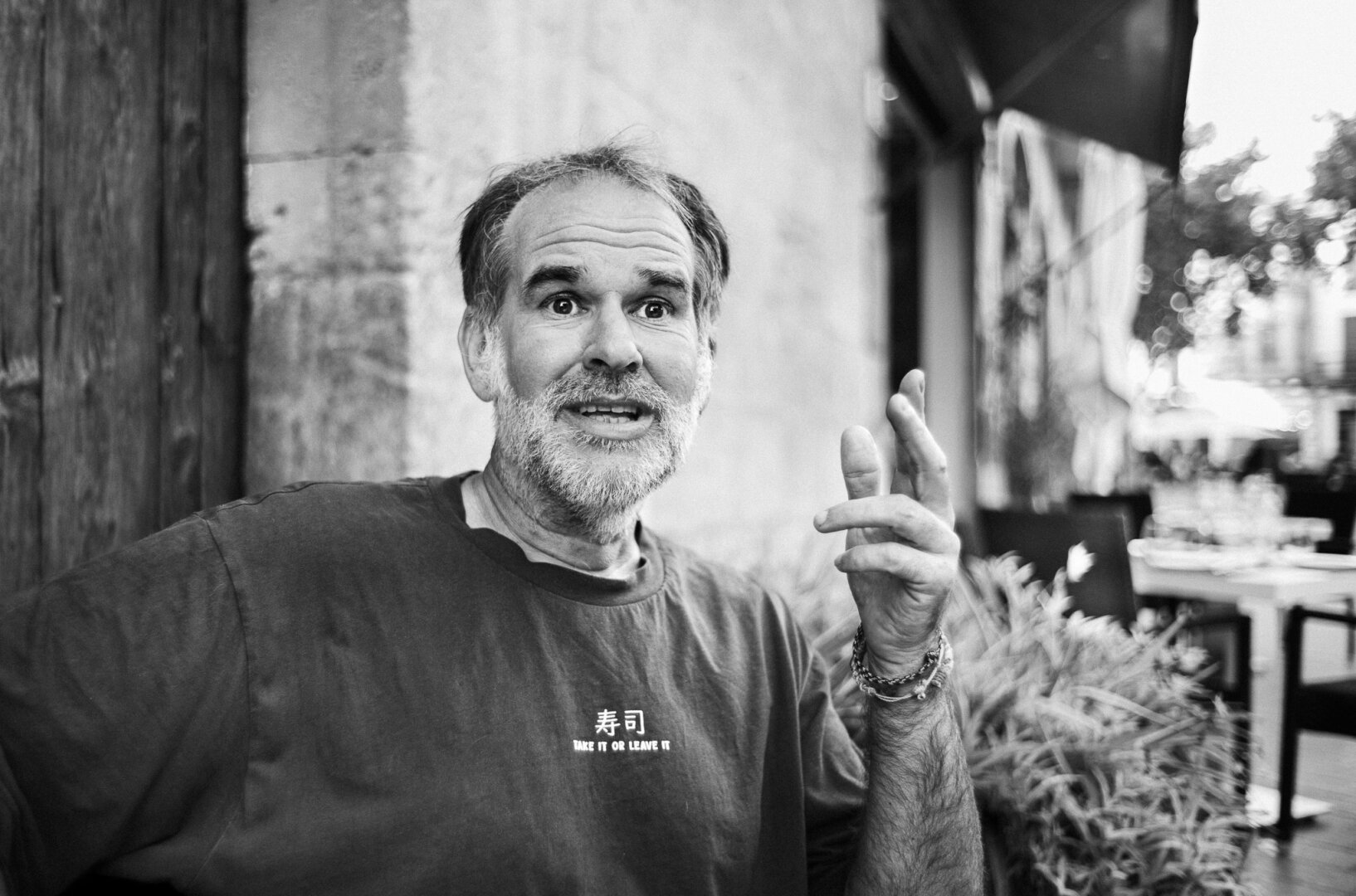 Black and white street portrait of a bearded man looking directly at the camera while gesturing with one hand, seated beside an outdoor café area with the background out of focus.