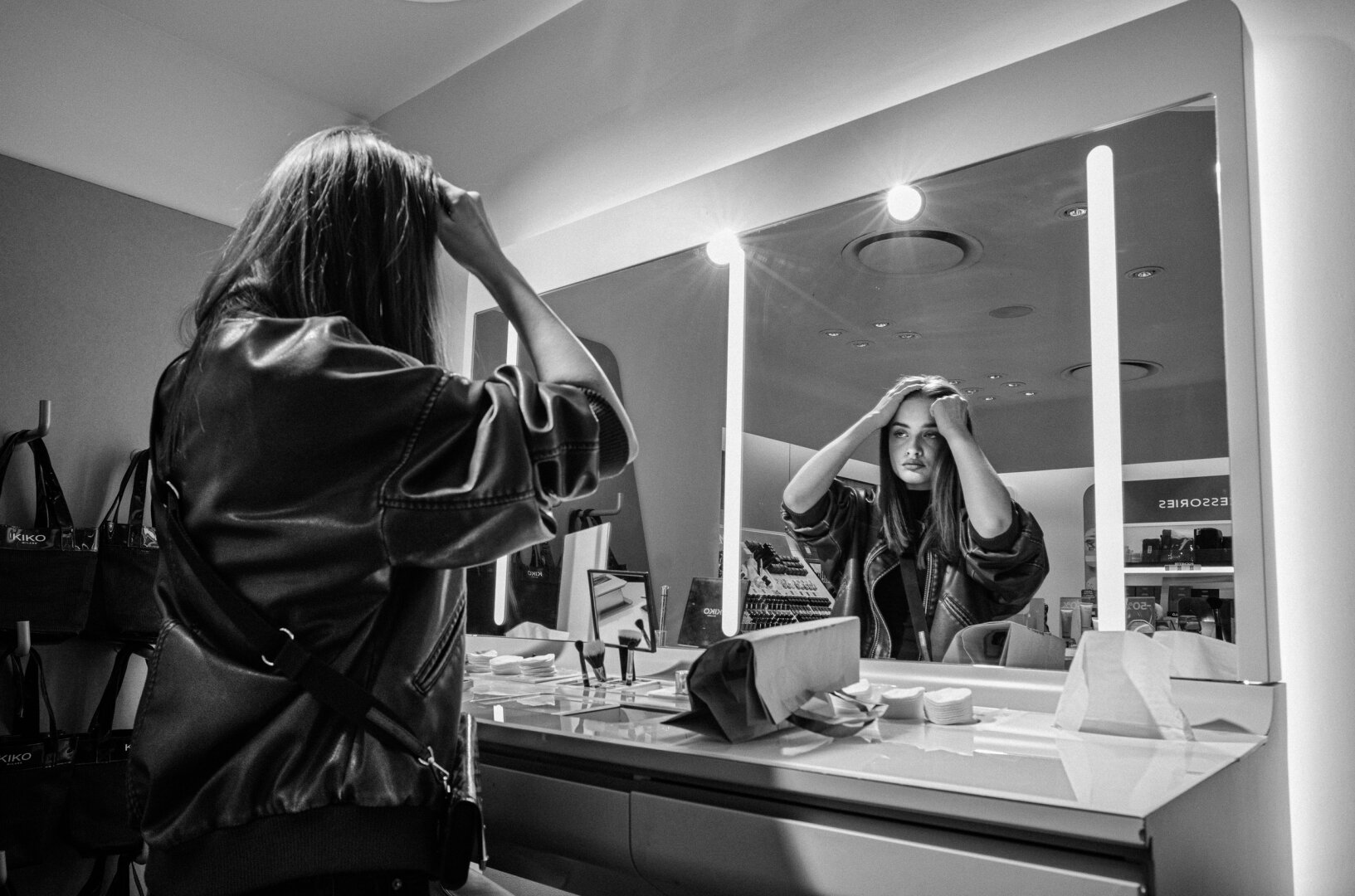 Black-and-white street photograph of a woman standing before a brightly lit cosmetics-store mirror, seen from behind and in reflection, with both hands raised to her head.