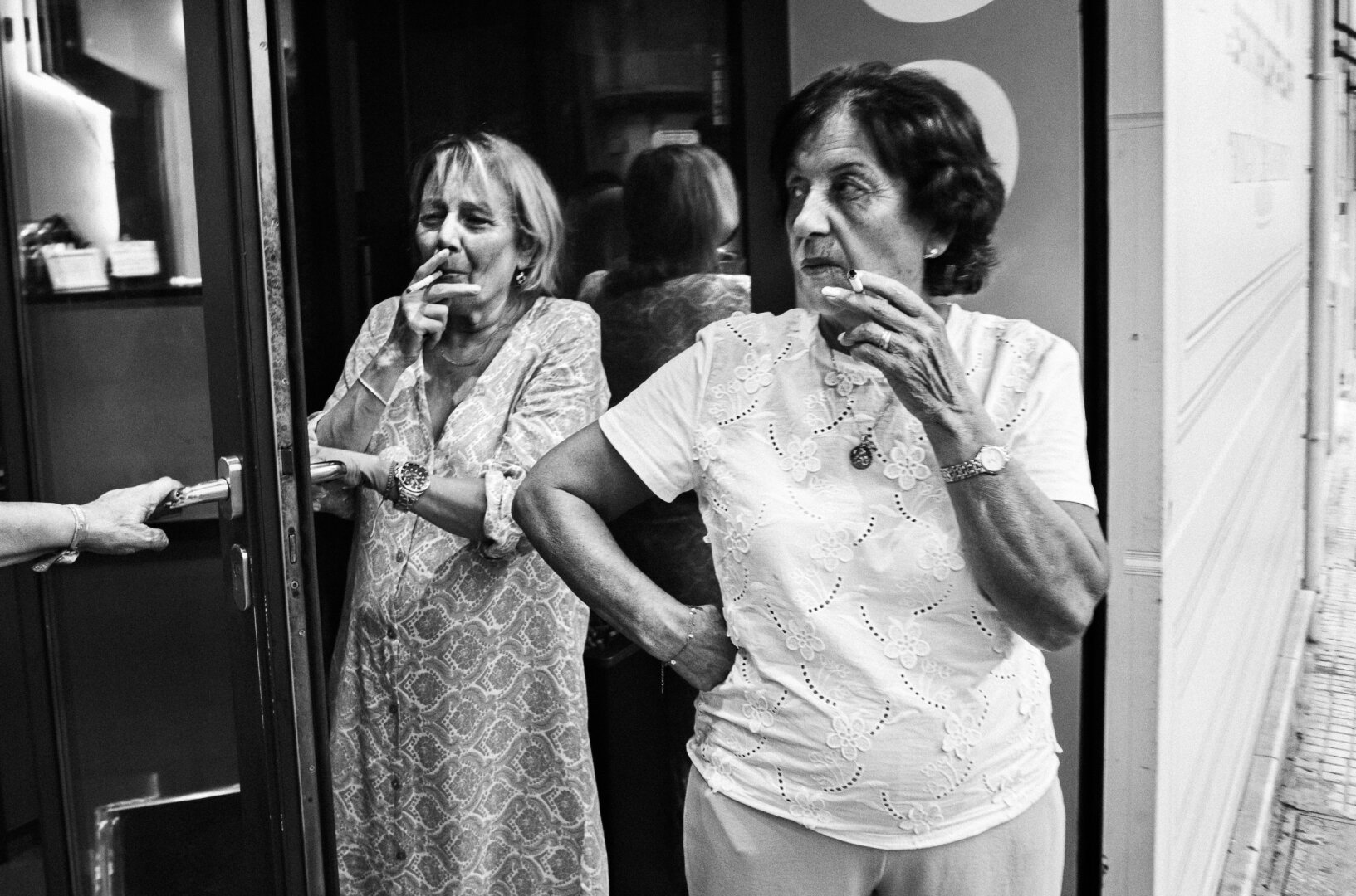 Black and white street photograph of two women smoking outside a bingo hall, one standing in the doorway and the other in the foreground, while a third person’s hand holds the door open.