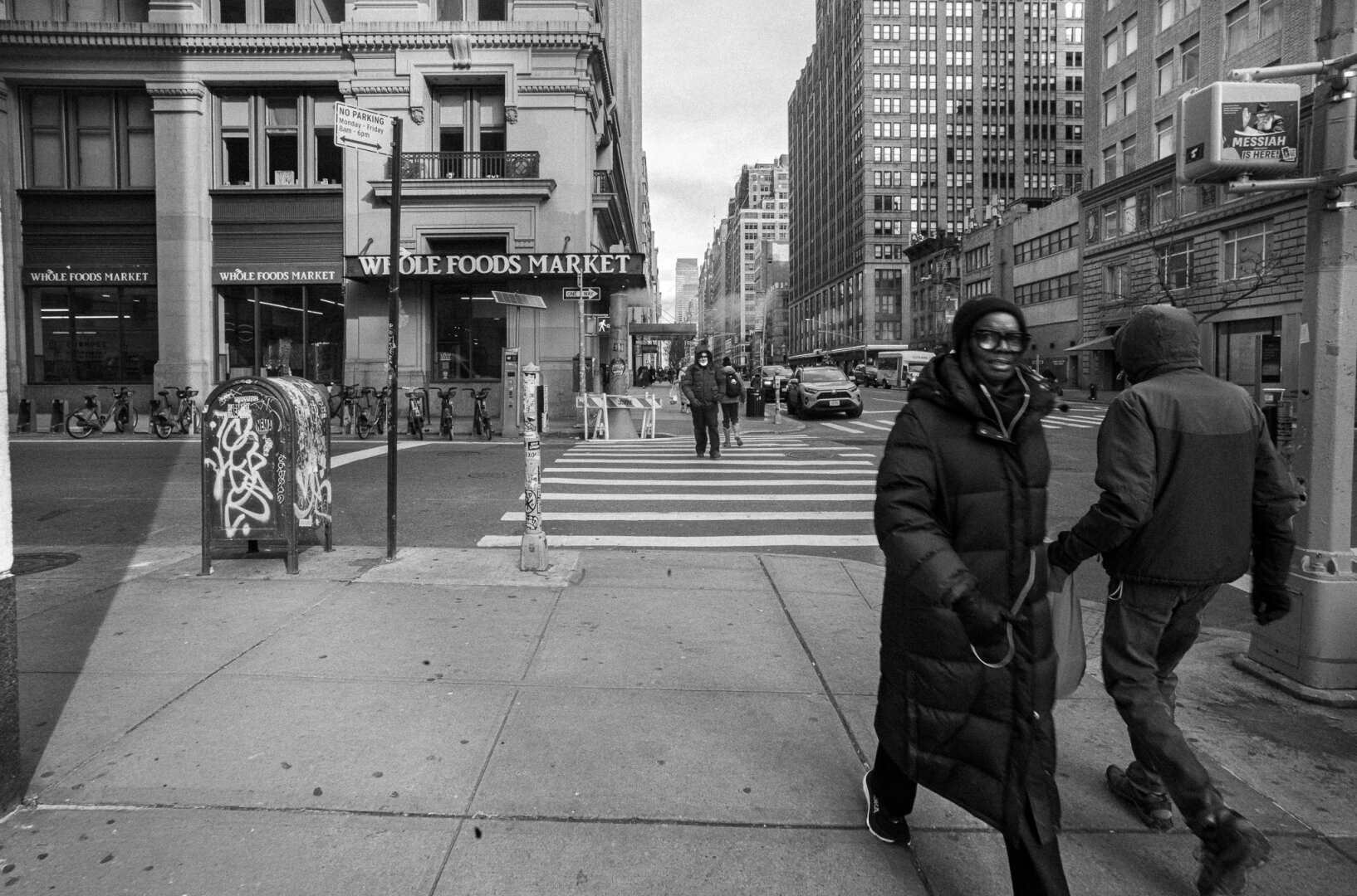 Black-and-white street photograph of two bundled pedestrians passing close to the camera at a city intersection, while another person waits across a striped crosswalk. Buildings, traffic and street signs fill the background.
