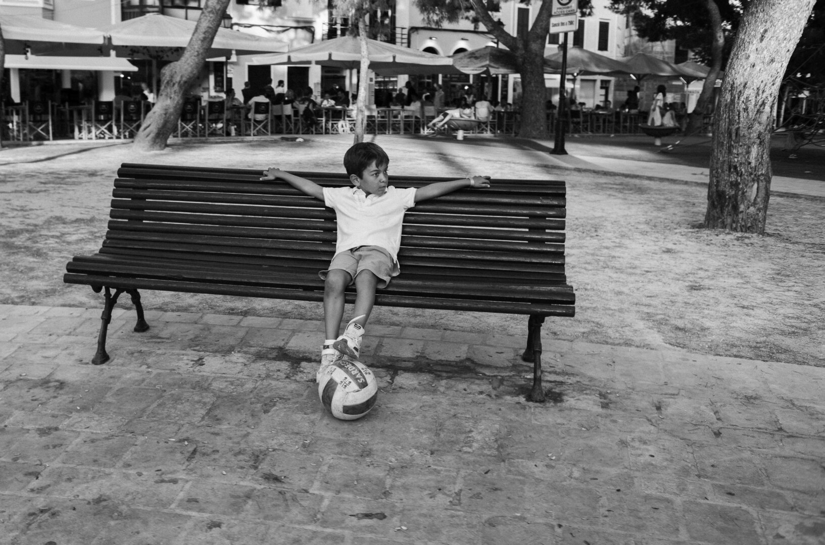 Black and white street photograph of a boy sitting alone on a long park bench, with his arms stretched across the backrest and one foot resting on a football. He looks to one side, with trees and café terraces in the background.