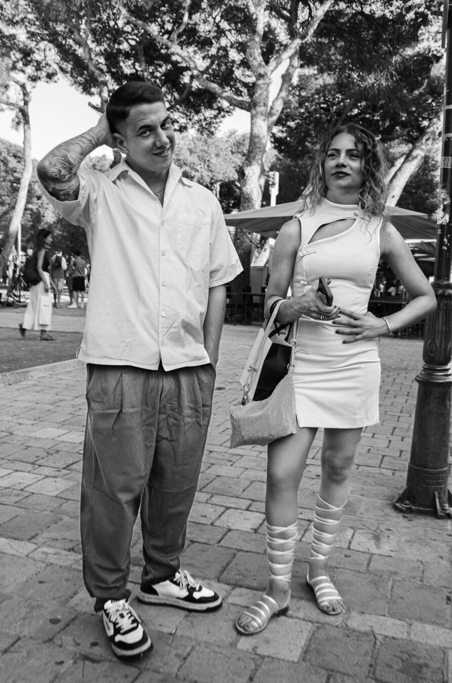 Black-and-white street photograph of a young man and woman standing side by side in a tree-lined public square. He looks at the camera with one hand behind his head, while she holds a phone and looks outside the frame.