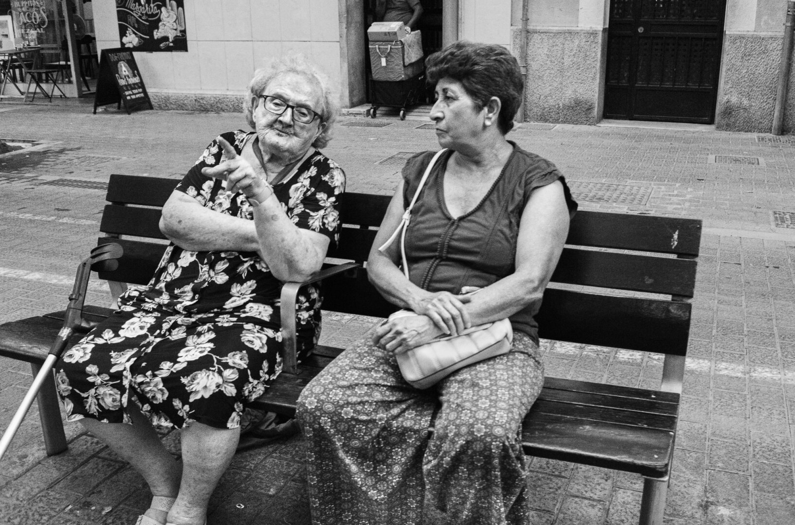 Black and white street photograph of two women seated on a public bench, one raising two fingers as she speaks while the other watches her, with a walking cane leaning against the bench.