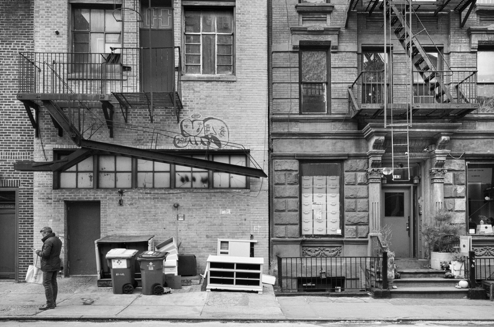 Black and white street photograph of a lone person standing at the edge of a sidewalk beneath a dense façade of brickwork, windows, railings and fire escapes.
