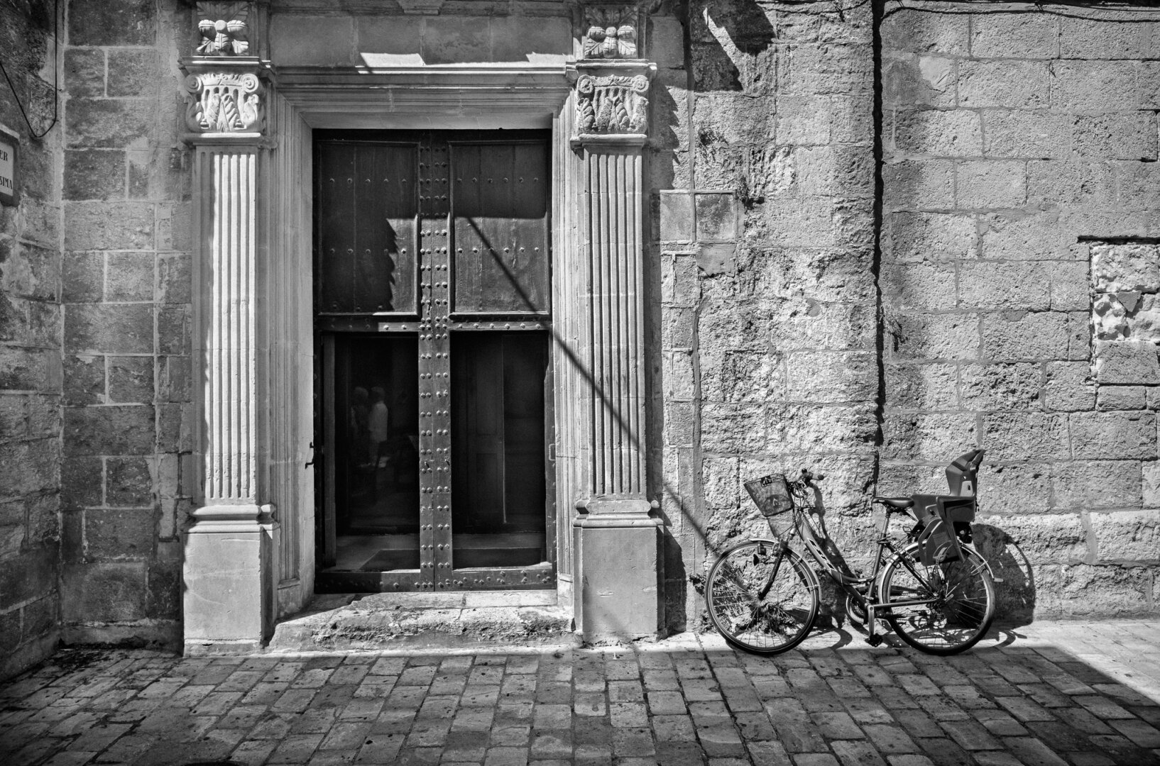 Black-and-white photograph of an ornate stone doorway in hard sunlight. A bicycle fitted with a child seat rests against the rough wall on the right, while deep shadows cross the door and pavement.