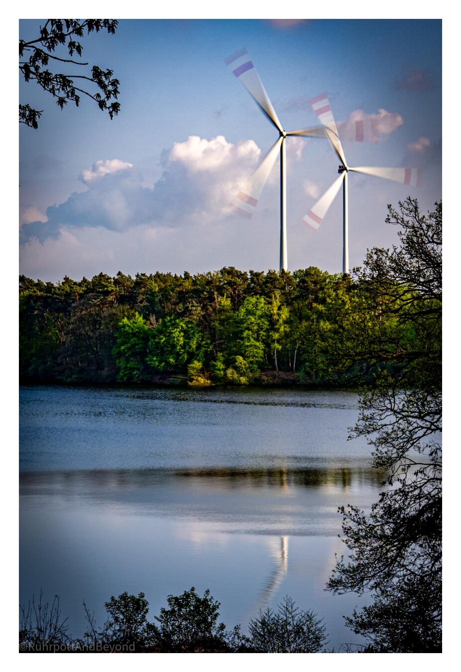 Im Vordergrund ein See bei Abendlicht. Im Hintergrund sieht man ein Waldstück und dahinter zwei gewaltige Windräder in Bewegung. Die Belichtungszeit wurde so gewählt, dass die Rotorblätter unscharf werden.