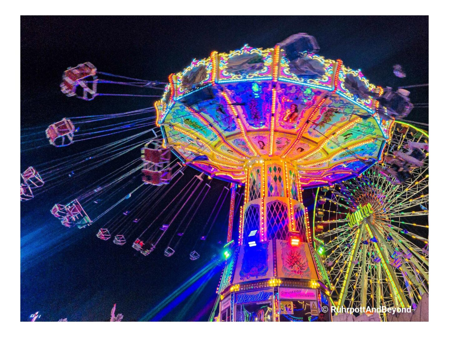 Ein farbenfroh erleuchtetes Kettenkarussell und ein Riesenrad bei Nacht.