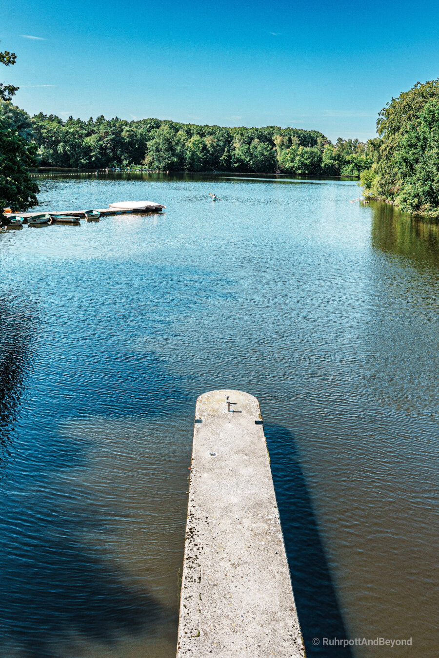 Blick über einen Betonsteg eines Wehres auf einen See, umrahmt von Bäumen, auf einen kleines Kajak mit Paddler. Strahlend blauer Himmel.