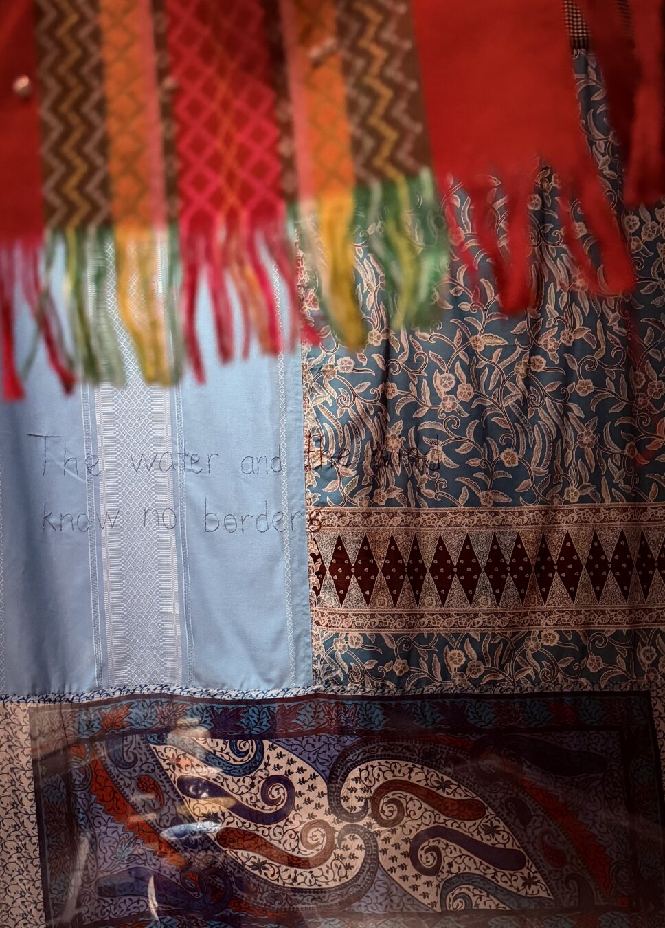 A close-up photograph of a textile art exhibition at the SMK museum. Colorful patterned fabrics hang layered together, including a bright red fringed scarf at the top, an intricate blue and brown floral batik pattern on the right, and a light blue embroidered fabric on the left that reads, "The water and... know no border".