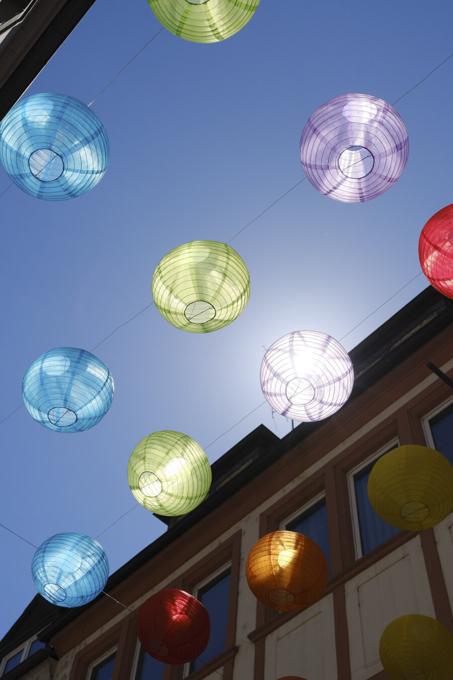 Colorful paper lanterns in blue, green, purple, red, yellow, and orange hang against a clear blue sky, with buildings visible below.