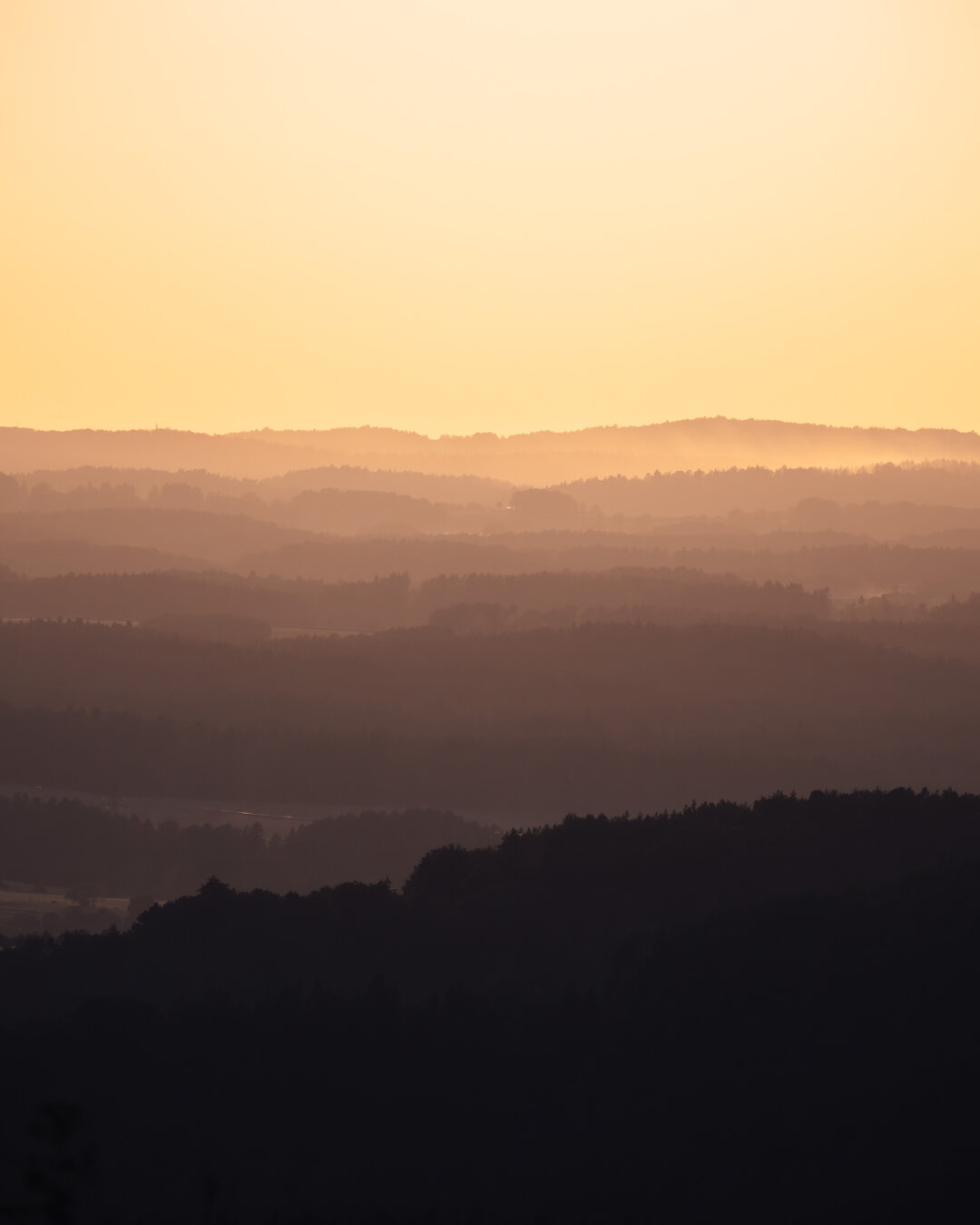Layered silhouettes of rolling hills fading into mist under a vast orange and yellow gradient sky.