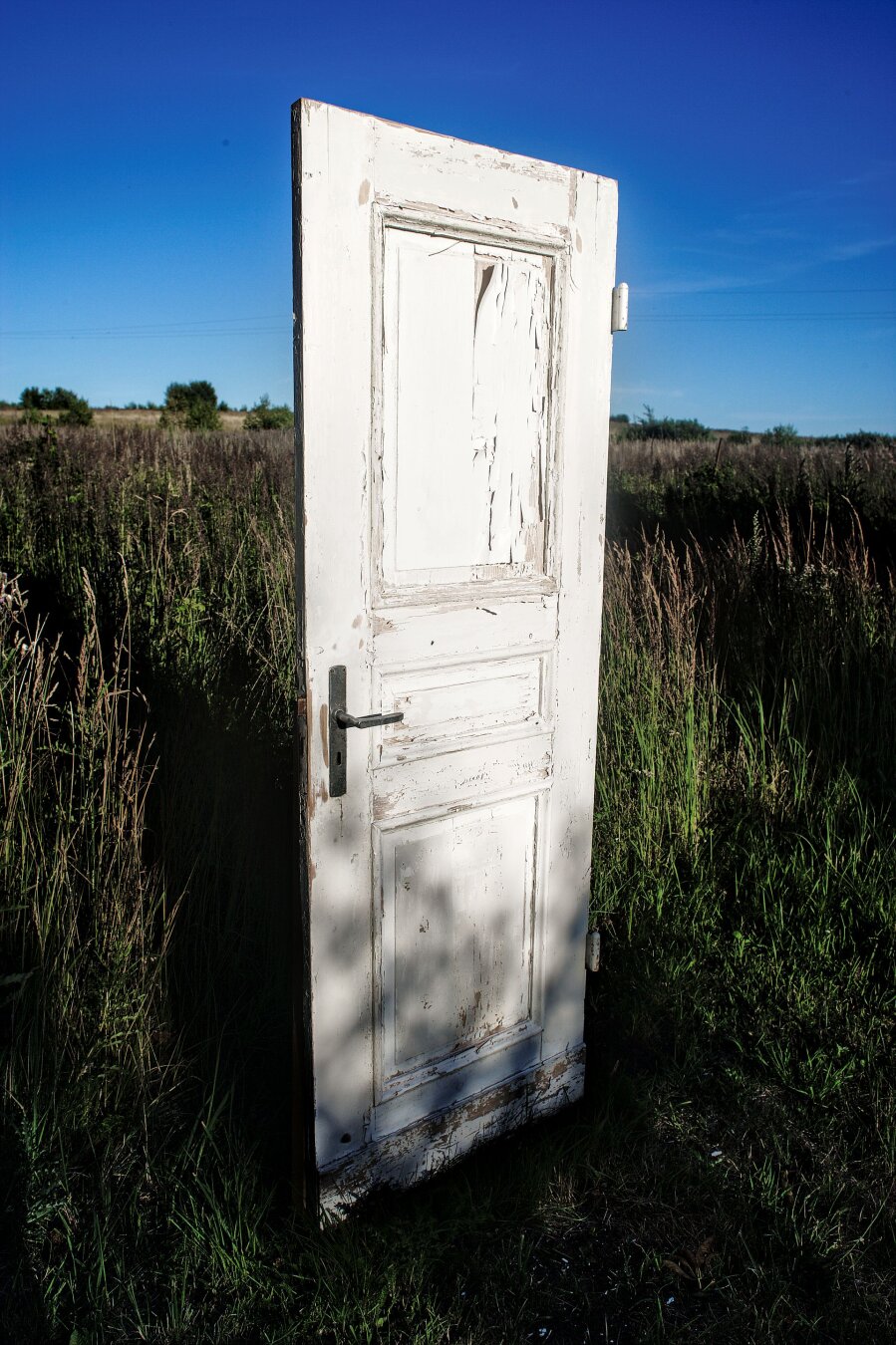Vertical medium-shot of an old, weathered white wooden door standing upright in the middle of a wild, grassy field in Janowo. The white paint on the door is peeling and cracked, revealing dark wood underneath. The door has a dark metal handle. The background shows a vast, open meadow with tall grasses under a clear, deep blue sky. The lighting is soft and directional, casting distinct shadows on the lower part of the door.