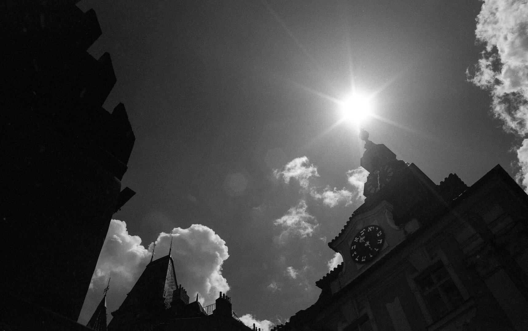 A high-contrast black and white photograph looking up at historic buildings in Prague. On the right, the Jewish Town Hall stands out, featuring its famous clock with Hebrew numerals. The sun is positioned directly above the tower's spire, creating a brilliant, sharp starburst effect. The building on the left is cast in deep shadow, forming a dark, geometric silhouette against a dark sky dotted with bright, fluffy clouds.