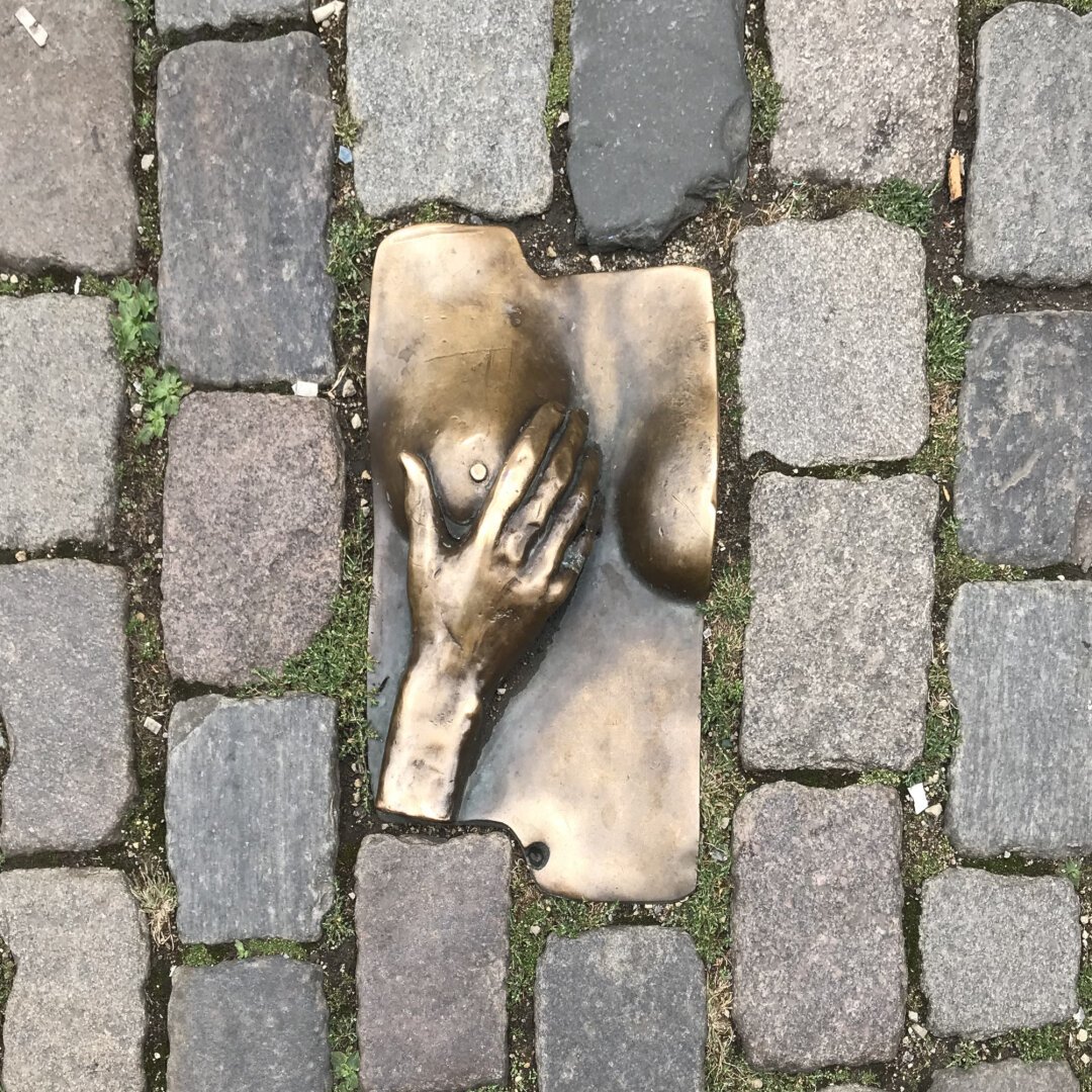 Bas-relief en bronze représentant un buste. Une main droite est posée délicatement sur le sein droit et semble le caresser. La sculpture est enchâssée dans le pavement.