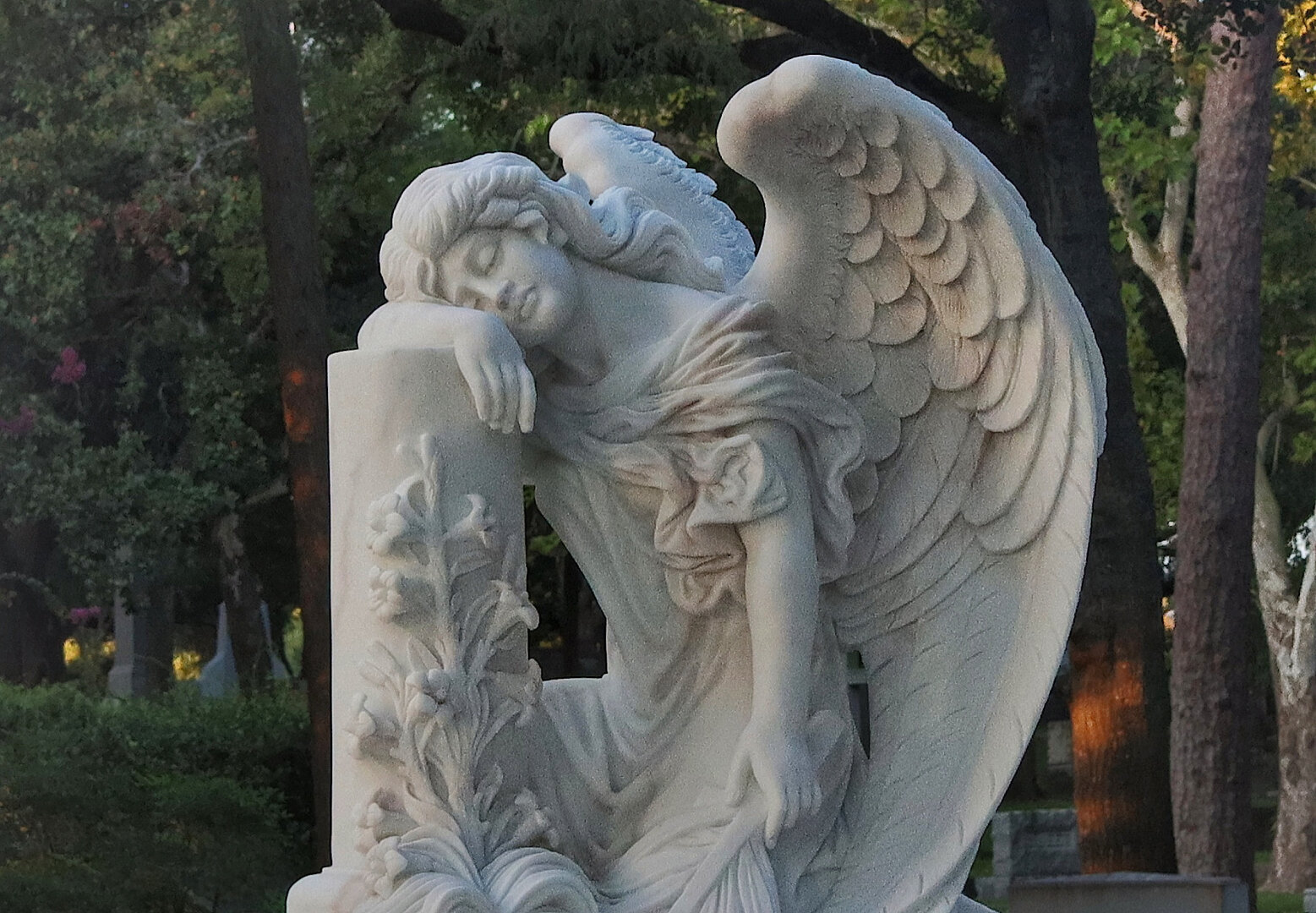 A beautiful marble angel stands by a pedestal and rests her arm and face upon it. She is pure white and surrounded by trees and grass in the shade of early morning. This is Glenwood Cemetery in Houston, Texas, USA.