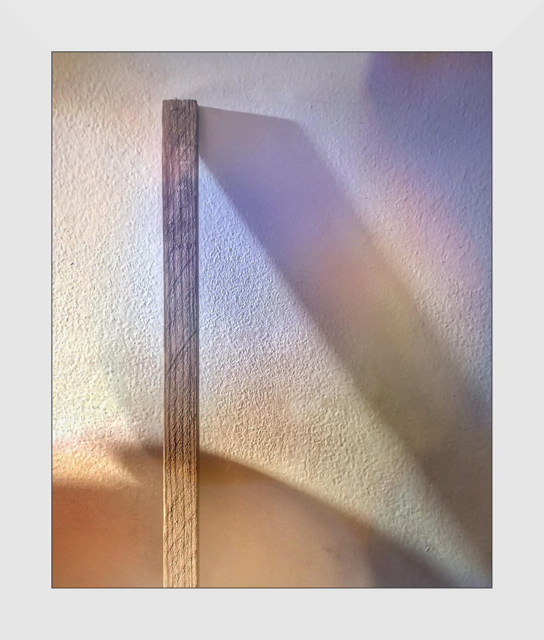 A stick of cut wood leans against a wall and casts a dramatic shadow to one side.