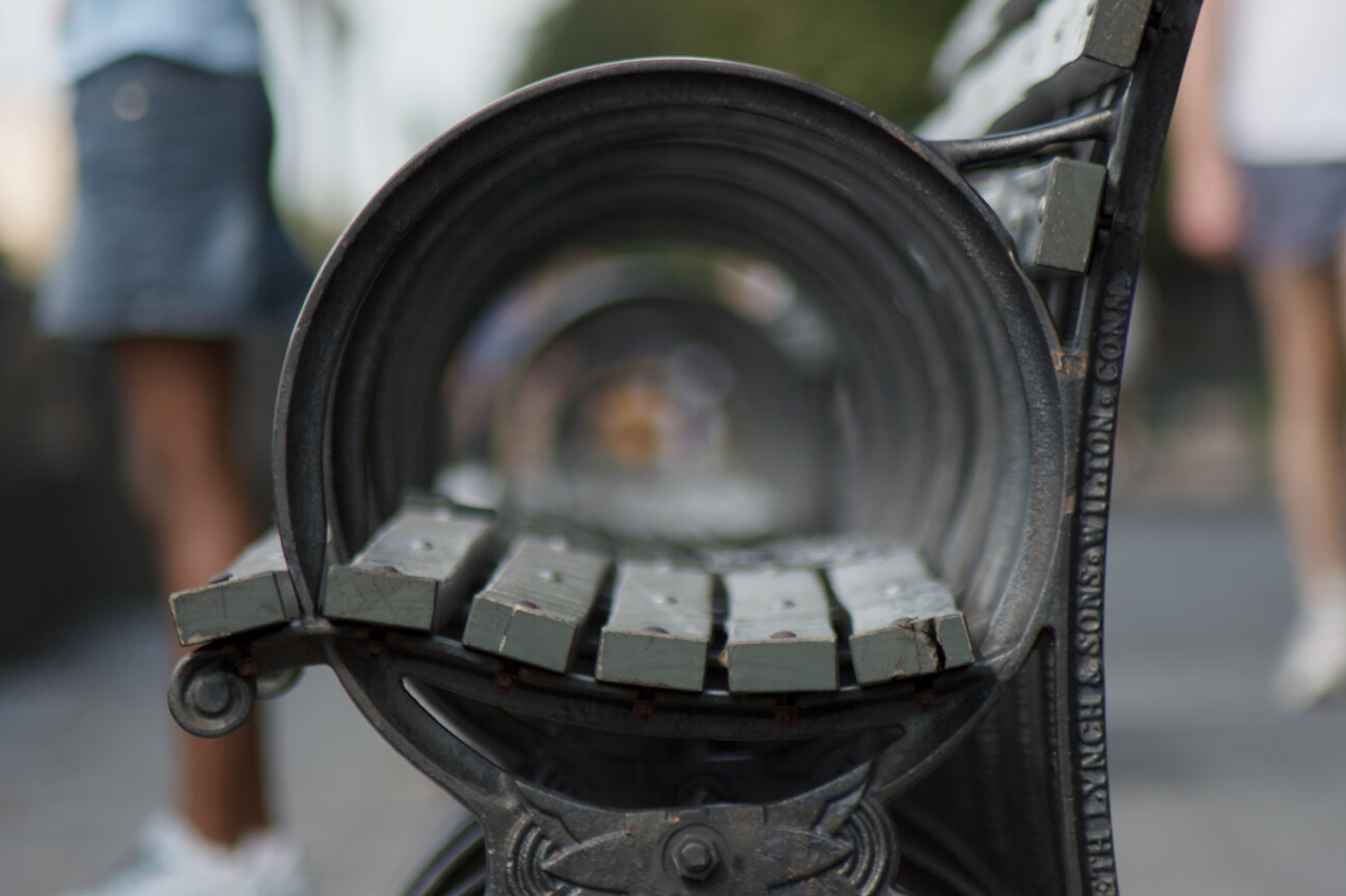 A very close photo of a typical New York park bench made of iron and wood. The picture is taken from the side, looking through the round iron armrest, so it makes a tunnel effect with other loops behind. The wooden seat parts are a little grey-green and blurry in the back. In the left and right background, you can see people legs standing and walking on the street, but they are out of focus.
