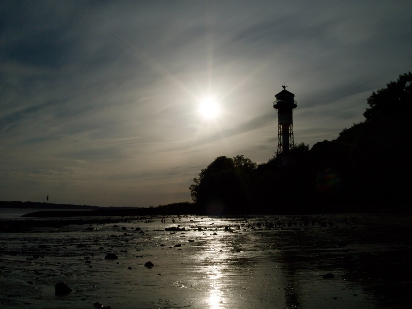 A lighthouse at the Elbe in Hamburg rises above the dark shoreline. Sunlight reflects on the wet mudflats at low tide, creating a scene of light, shadow and stillness.