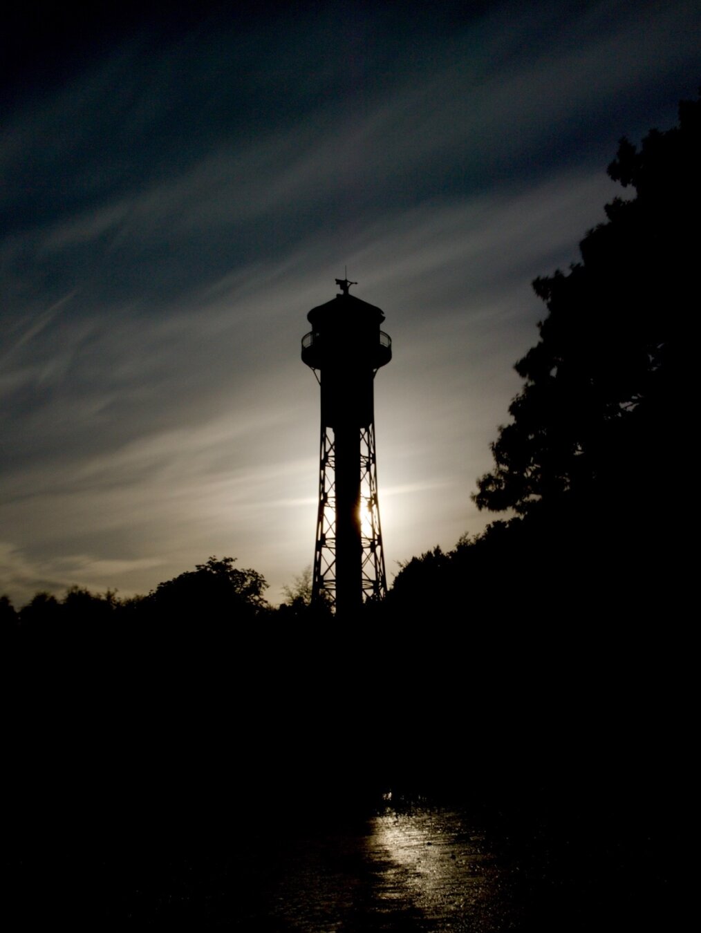 Silhouette of a lighthouse tower with metal structure in the evening. Dark clouds in the sky behind it. Dark trees and plants are on the right side. In front is mudflat with a small light reflection. turen und kleine Unregelmäßigkeiten.