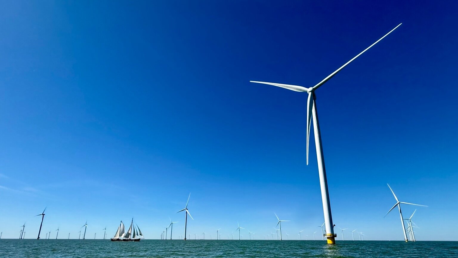 Low-angle perspective of an offshore wind turbine against a deep blue sky, with multiple wind farms visible on the horizon. A traditional sailing vessel passes below the towering structure, connecting old maritime traditions with modern renewable energy infrastructure.
