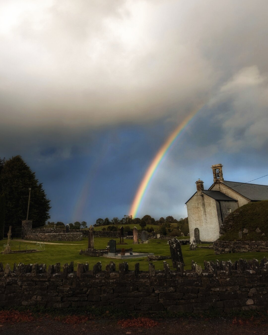 A partially cloudy sky with a double rainbow, and a small church on the right hand side with graveyard. A stone wall in the foreground