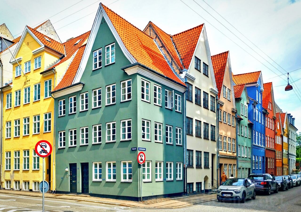 Fila de edifícios coloridos com telhados vermelhos inclinados, janelas brancas, carros estacionados e sinais de trânsito numa rua urbana.