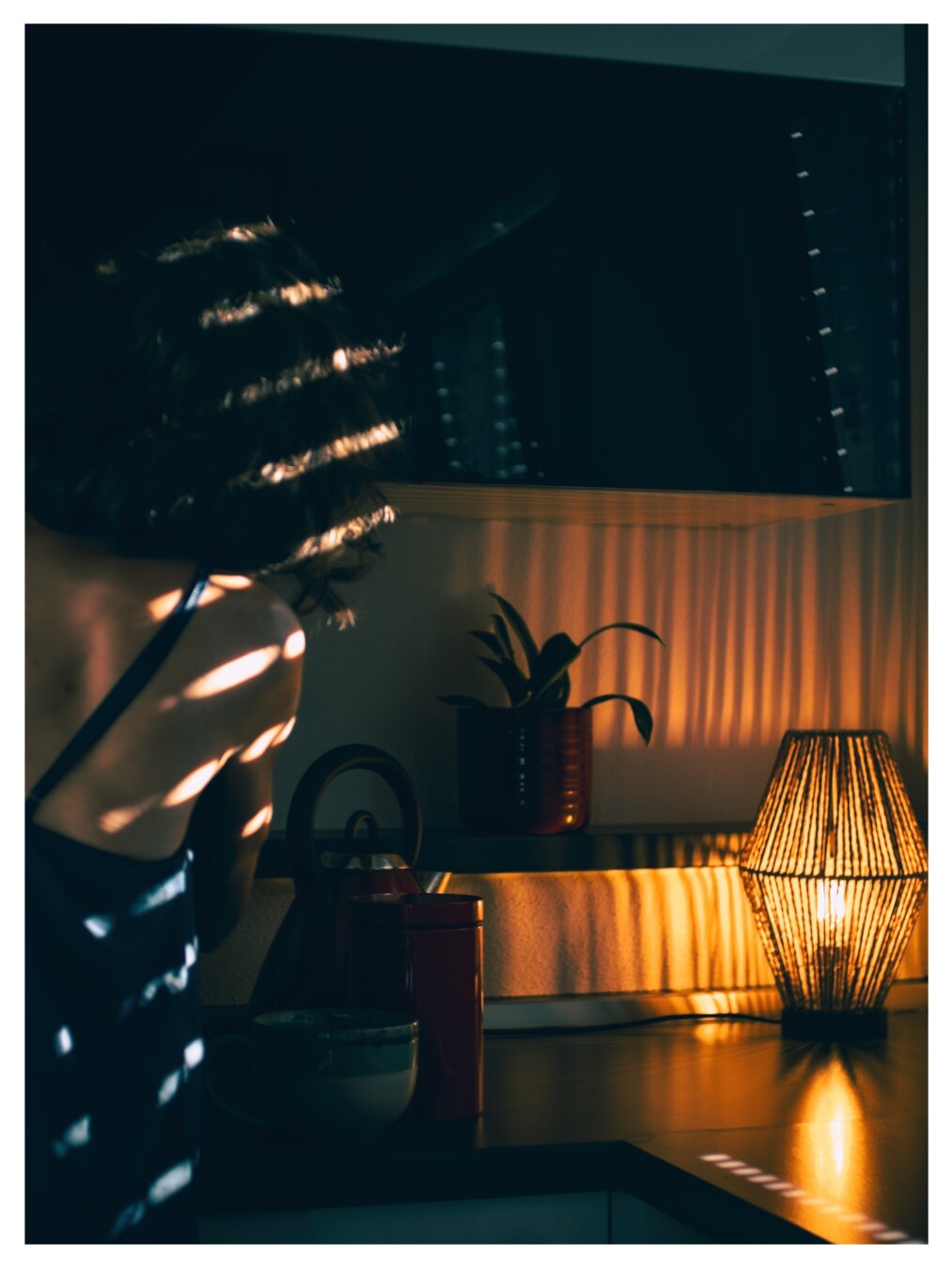 warmly lit interior scene. sunlight shines through blinds casting stripes across the back of a person standing in the foreground while a table lamp glows beside a potted plant on a countertop and creates a vertical pattern of shadow and light on the wall