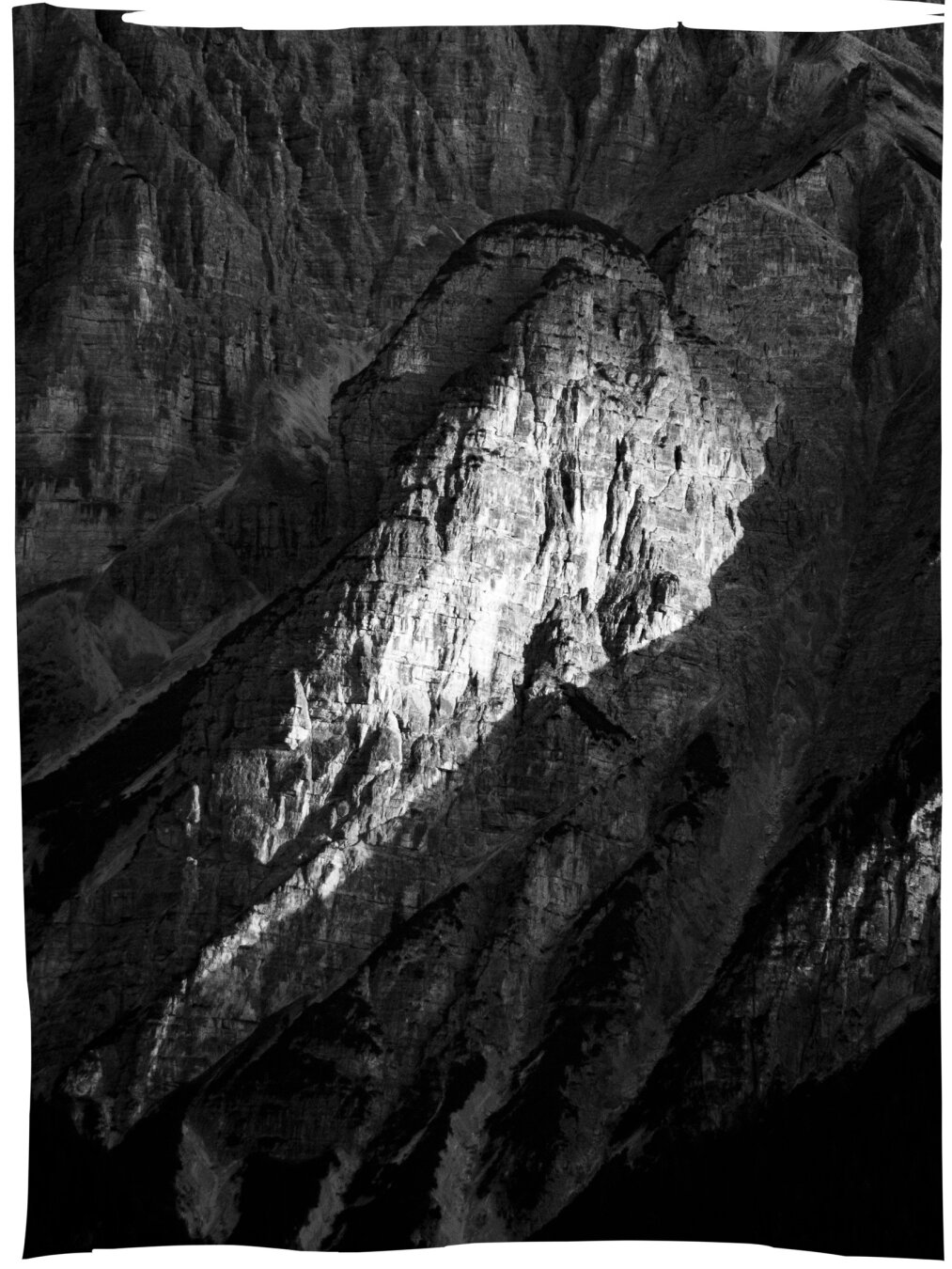 black-and-white photograph of a mountain ridge where a spot light on the rocks creates a focal point 
sunlight casts sharp shadows emphasizing the rugged textures and the contrast between light and dark