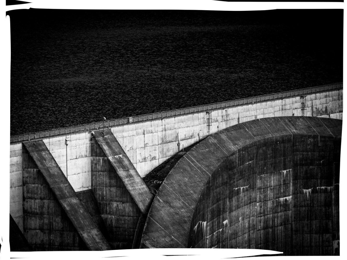 black-and-white photograph of a massive dam structure with concrete walls and geometric spillways
dark water above contrasts with the sharp angles and textures of the dam
architectural power and engineering precision shows off