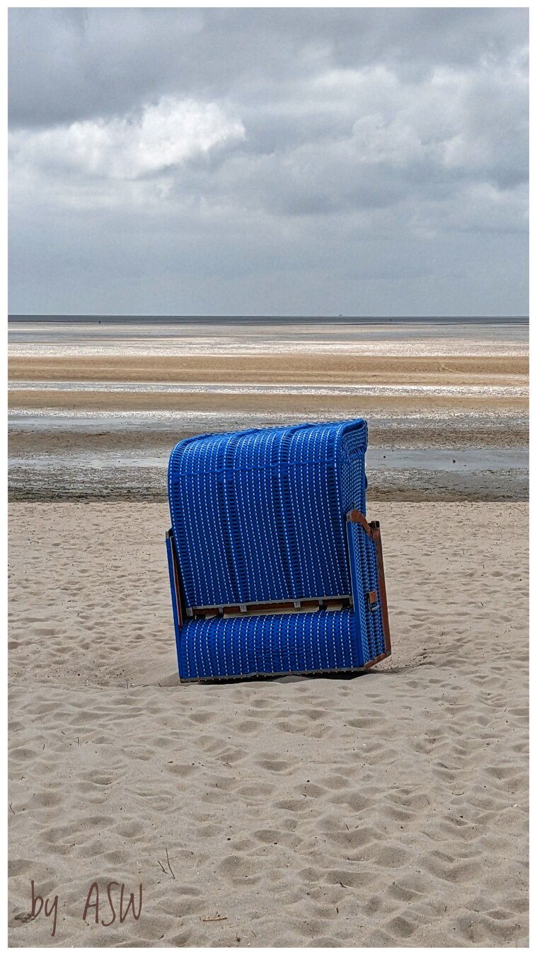 Ein blauer Strandkorb auf Sand, im Hintergrund das Meer / 
A blue beach chair on sand, with the sea in the background.