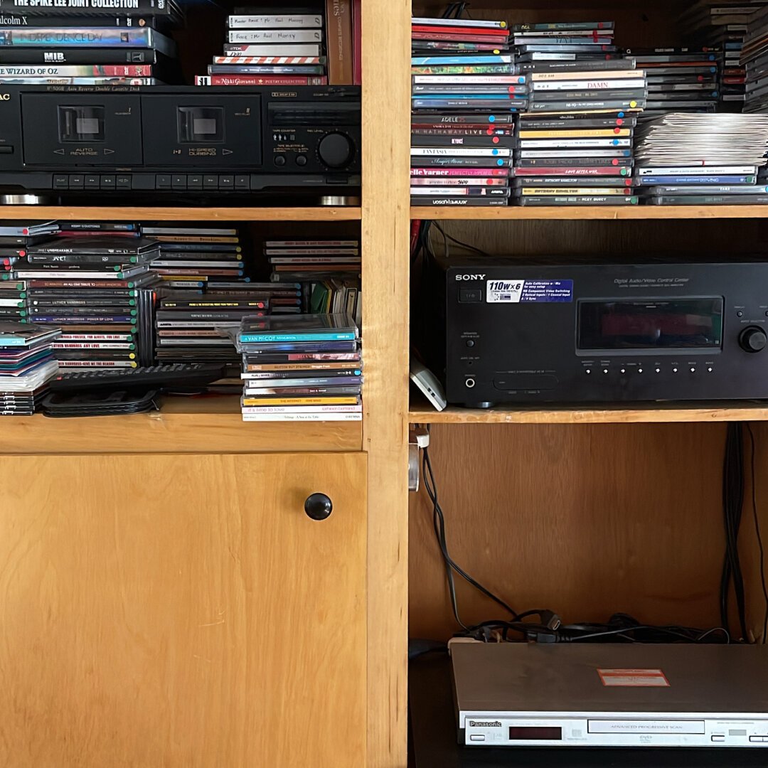 a circa 2004 home theater system amplifier/switcher, DVD player and 5 disc CD changer with a dual tape deck sitting in a birch wood cabinet surrounded by various CDs and cassette tapes. 