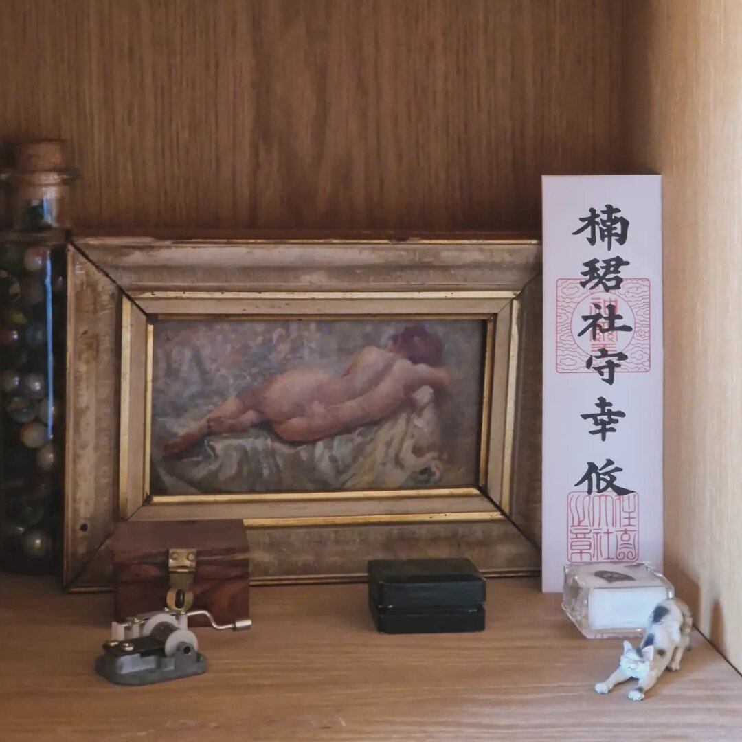 A quiet wooden shelf with a small framed reclining nude, a Japanese ofuda standing beside it, and several small collected objects: a tiny mechanical device, wooden and black boxes, a clear case, and a miniature stretching cat. The composition is warm, slightly faded and domestic, like a small cabinet of things gathered over time.