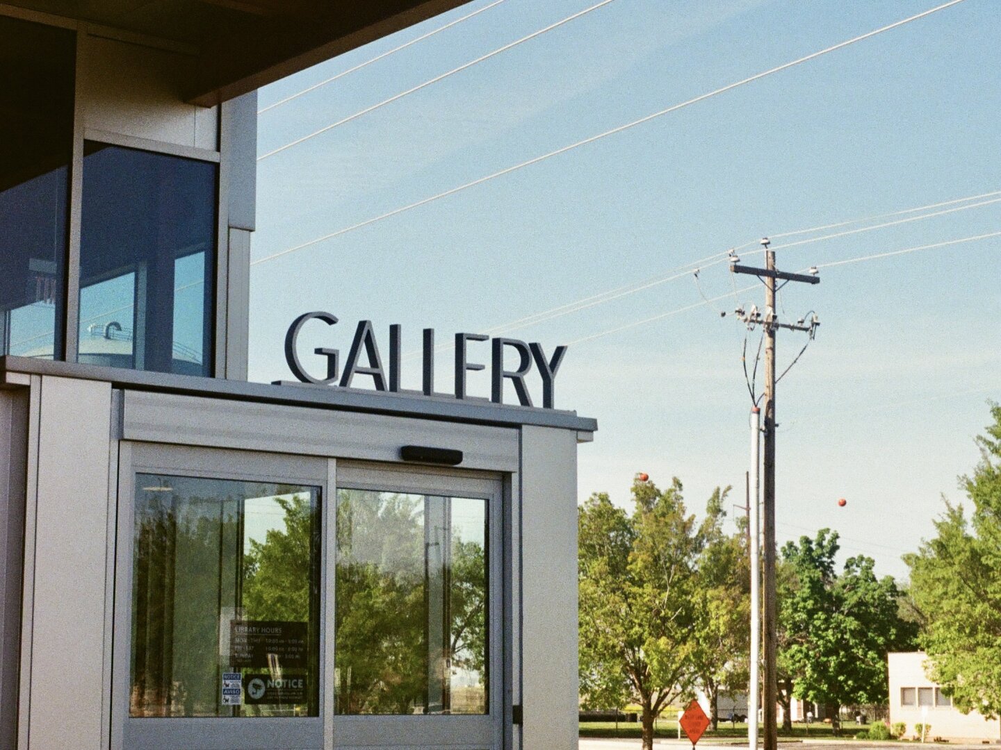 Lettering above a sliding glass door that reads GALLERY. A power pole and construction sign are visible among trees in the background under a clear blue sky.